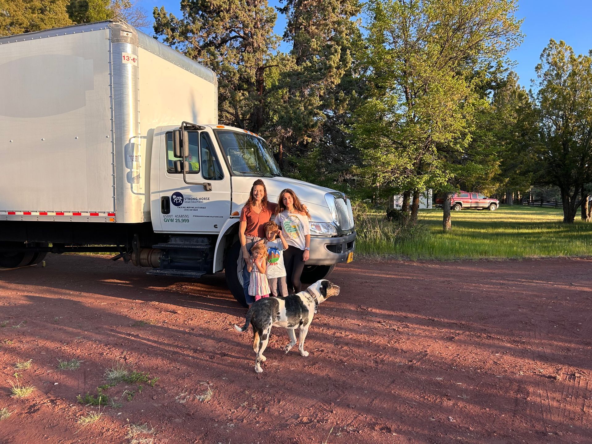 Woman with two dogs standing beside a white truck on a red dirt road by trees