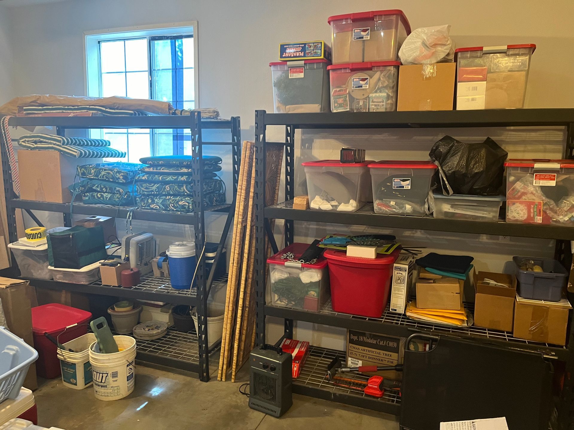 A cluttered storage room with metal shelves, plastic bins, boxes, and household supplies near a window.