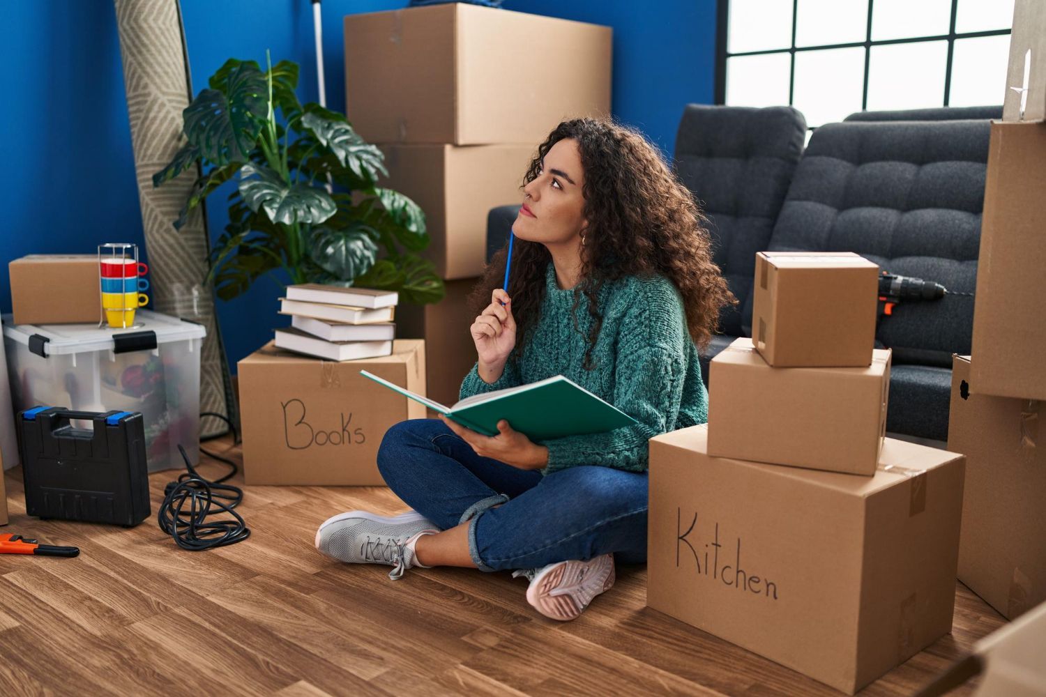 Woman sitting on the floor in a moving room, holding a notebook beside labeled boxes and packed belongings