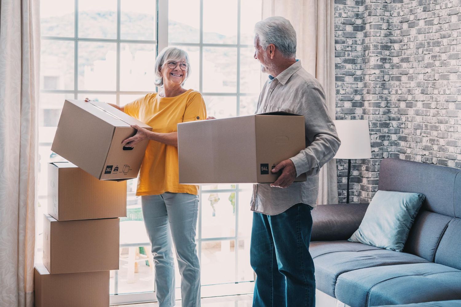 Two people holding moving boxes in a bright living room beside a blue couch