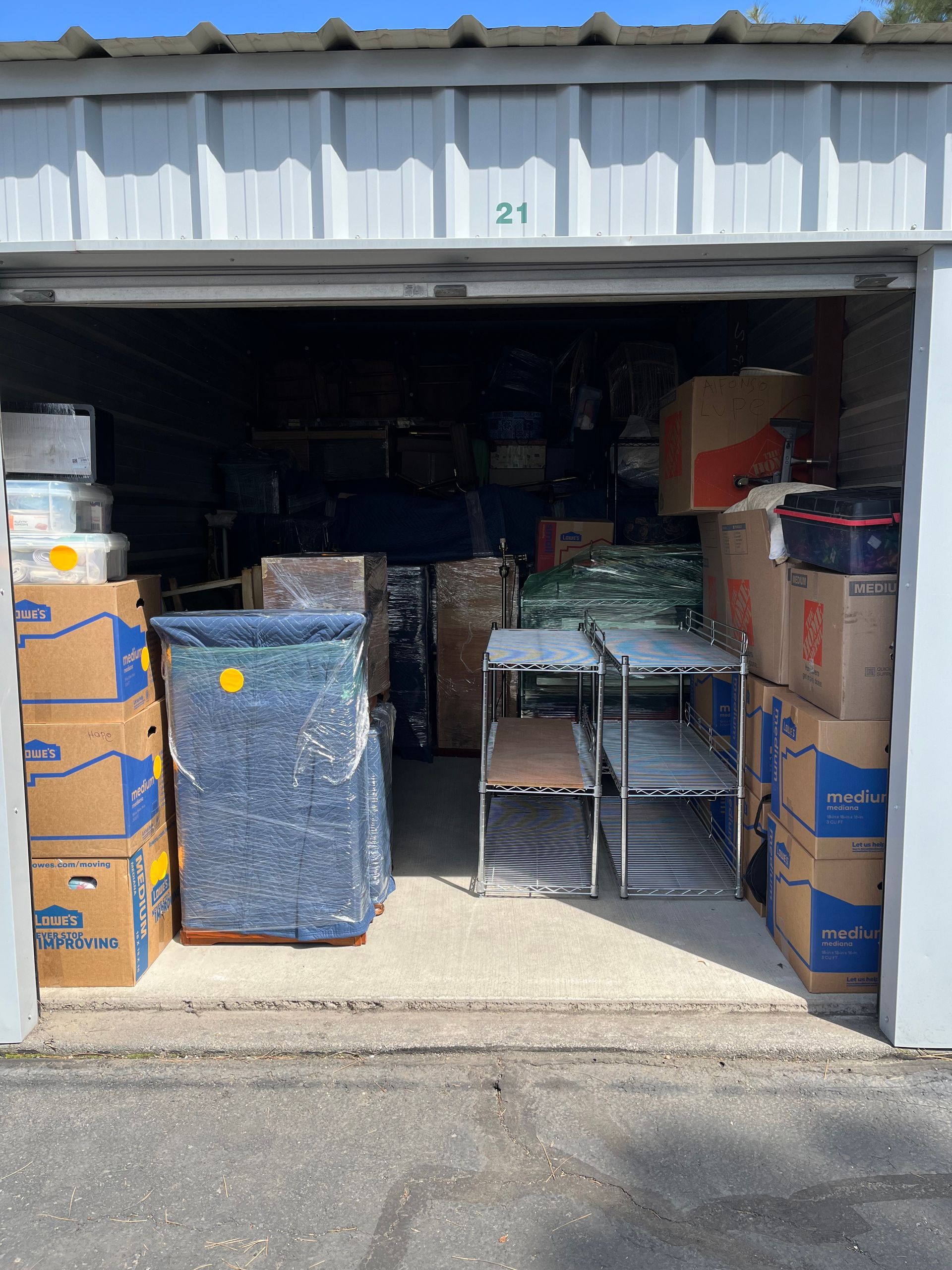 Open storage unit filled with stacked boxes, chairs, and wrapped furniture inside a metal doorway.