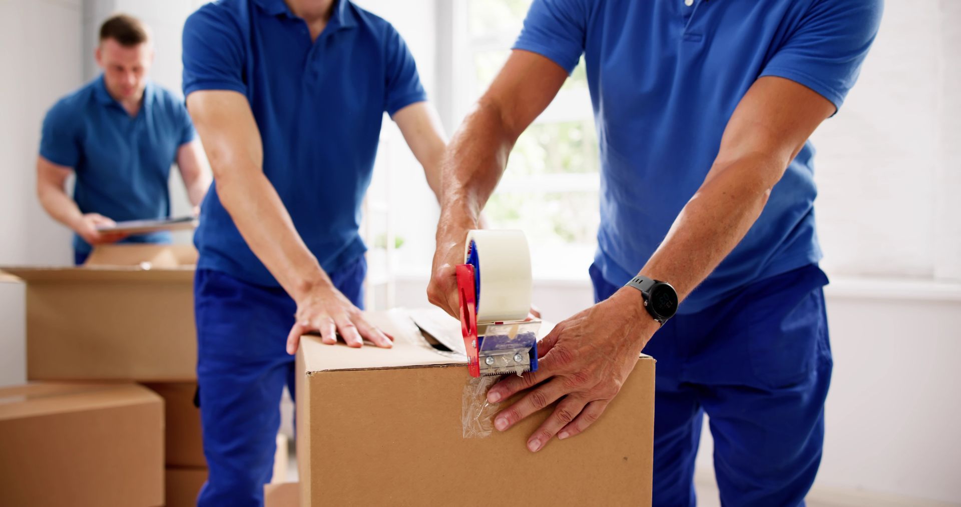 Movers packing cardboard boxes; one tapes a box, two others nearby in blue shirts.