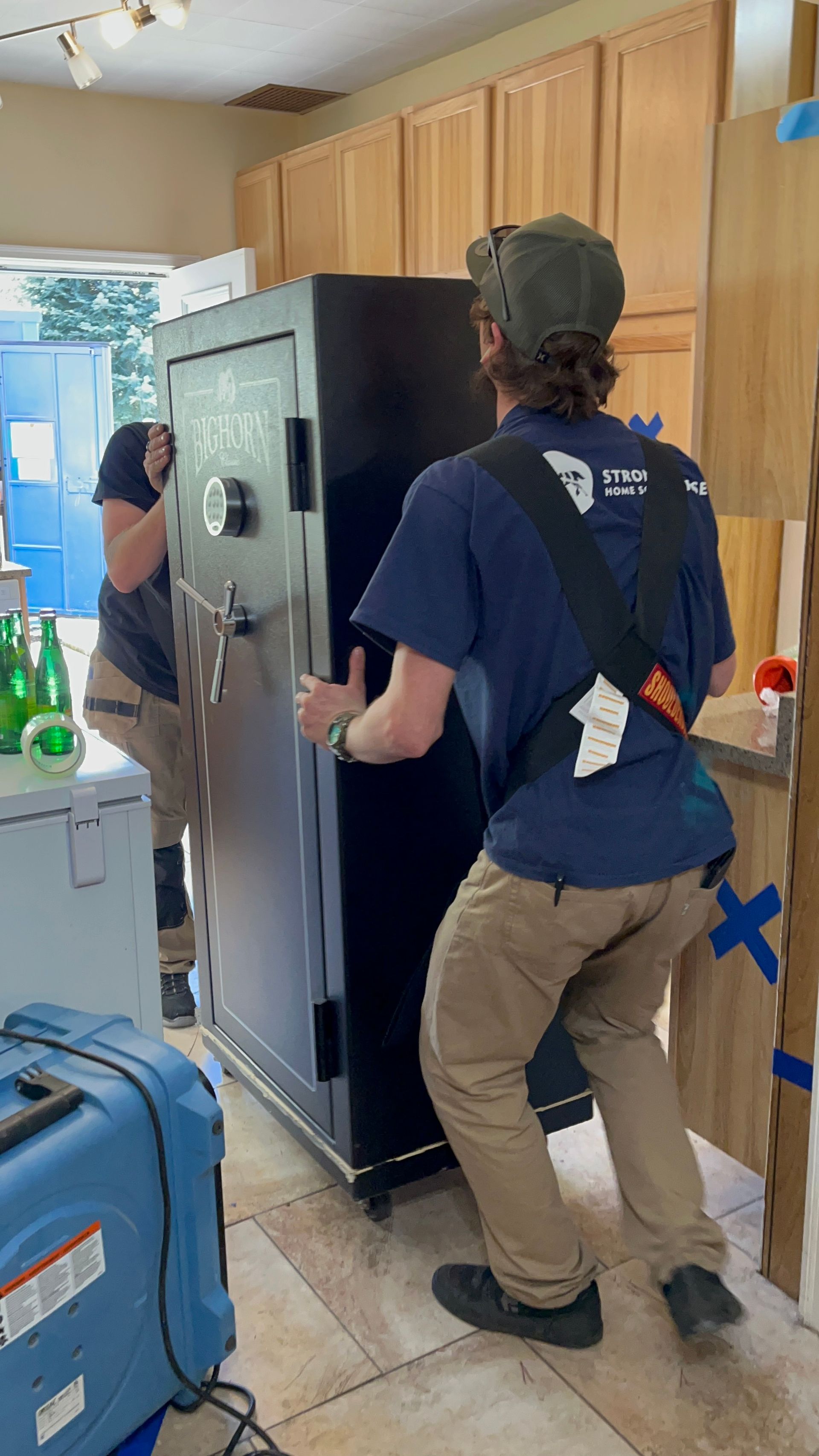 Person moving a large black appliance in a kitchen with wooden cabinets and a blue suitcase nearby