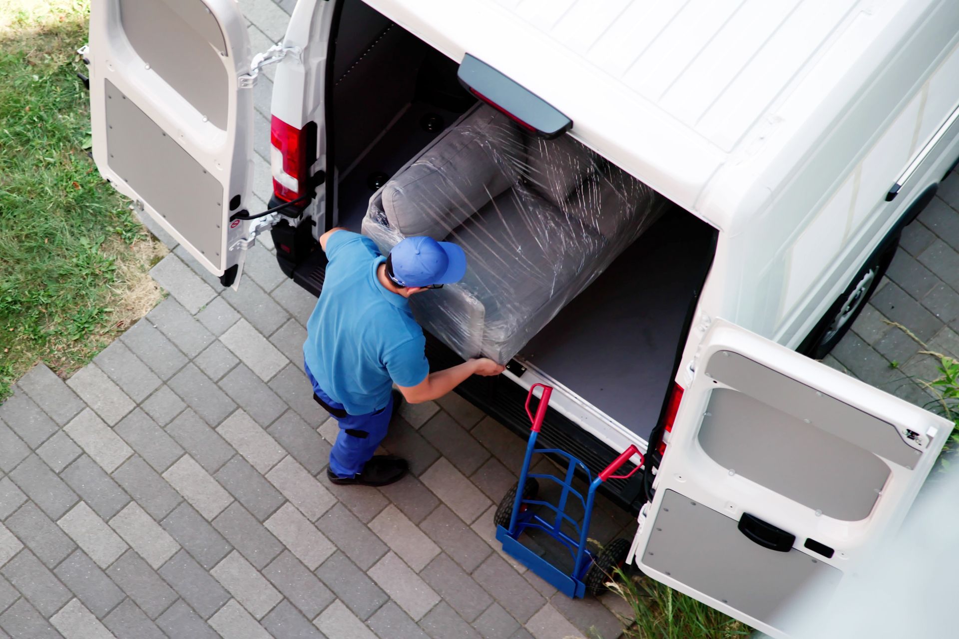 Man loading wrapped sofa into a white van from a brick patio. Blue shirt, hat, and a dolly are visible.