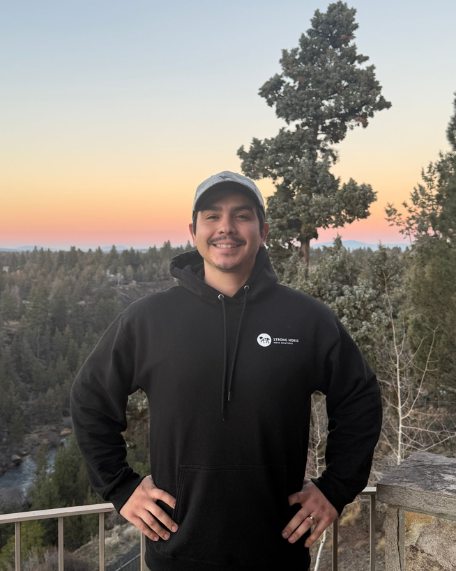 Man in black hoodie with hands on hips, standing outdoors with a sunset backdrop.