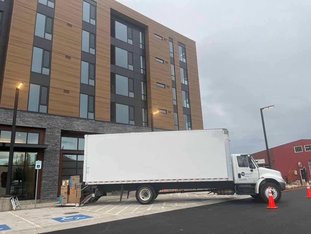 White delivery truck parked beside a brick apartment building on a city street with traffic cones.