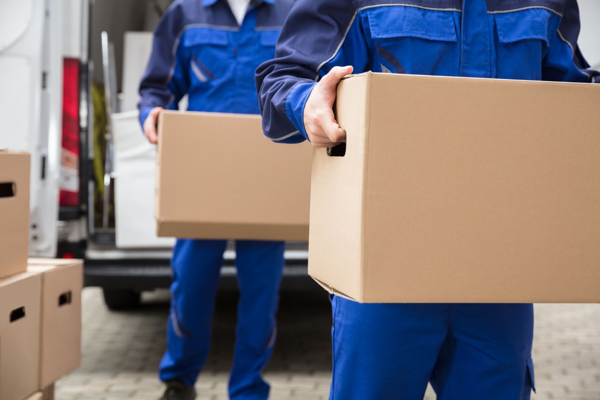 Two men in blue overalls are carrying cardboard boxes in front of a van.