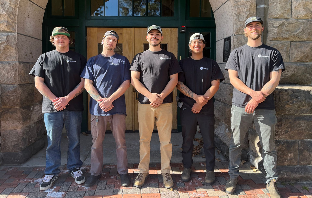 Five people standing in front of a stone archway entrance, wearing dark shirts and caps.