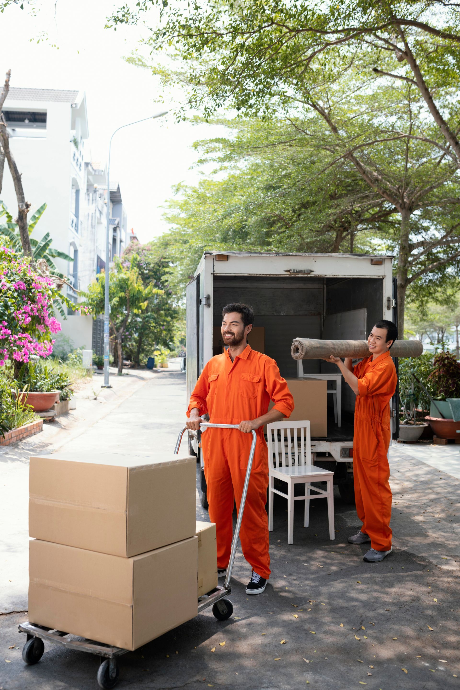 Two movers in orange jumpsuits load a truck. One pushes a cart with boxes; the other carries a rolled rug.