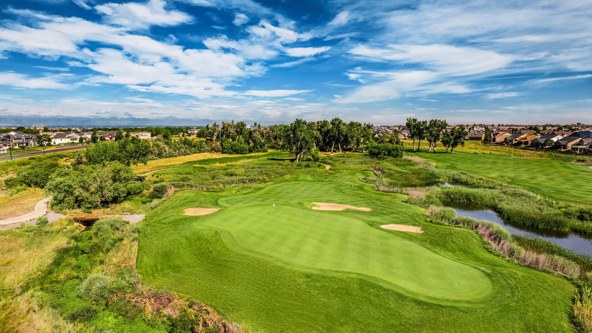 An elevated view of a bright green golf course fairway surrounded by trees, water, and suburban homes under a blue sky.