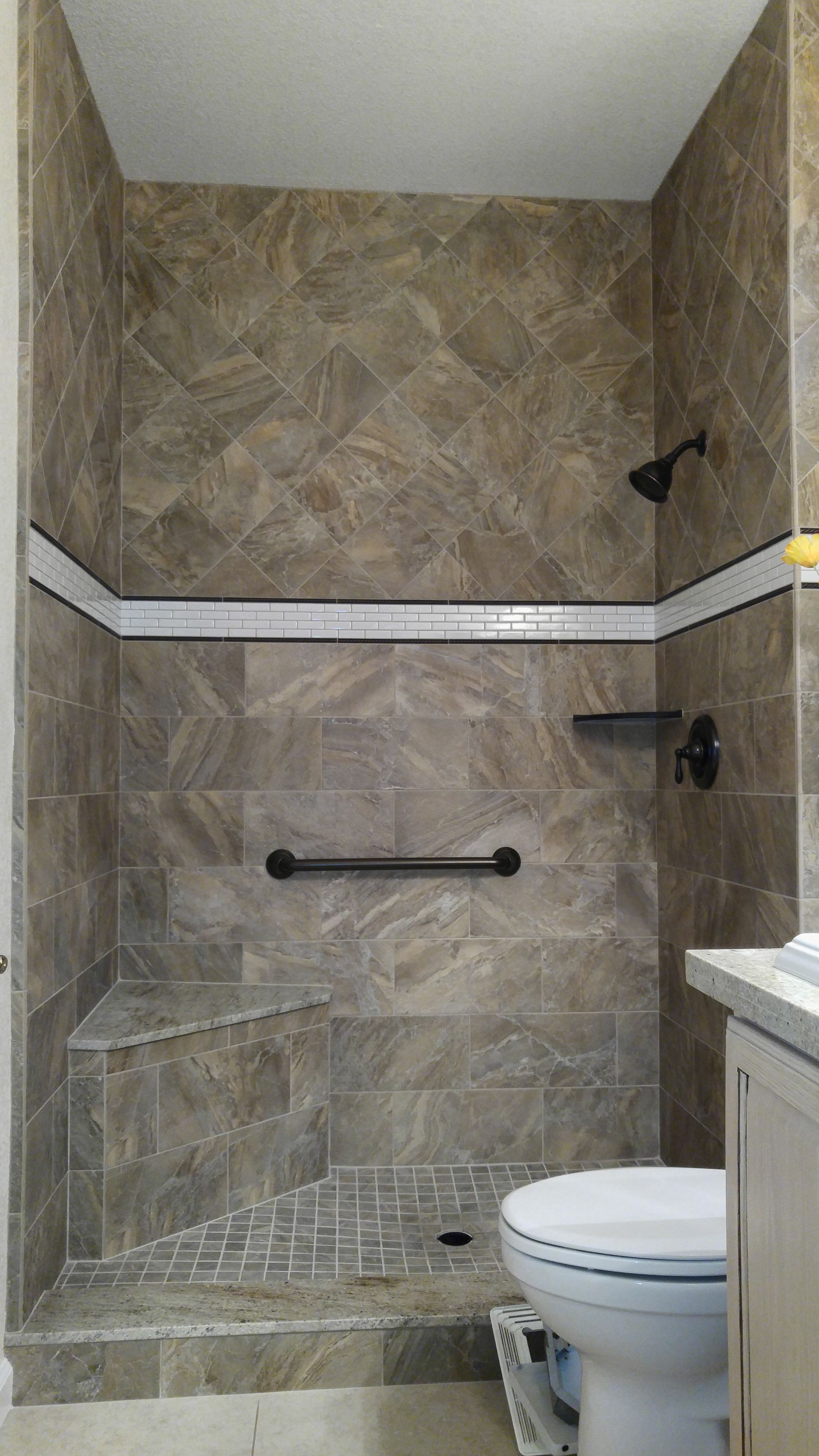 Tiled shower stall with bench, grab bar, and dark fixtures. Tan and brown tiles, white trim.
