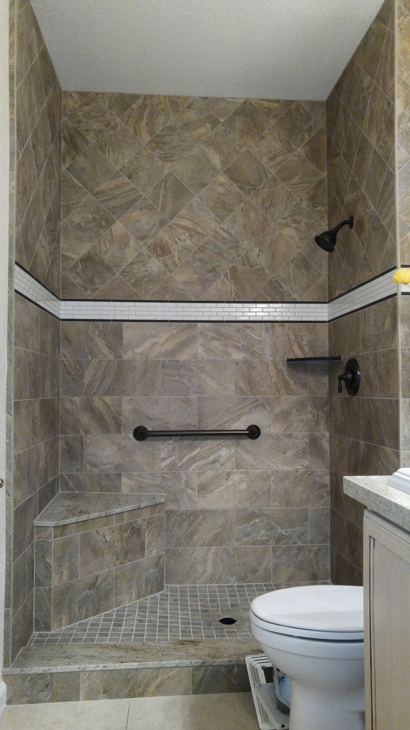 Tiled shower stall with bench, grab bar, and dark fixtures. Tan and brown tiles, white trim.