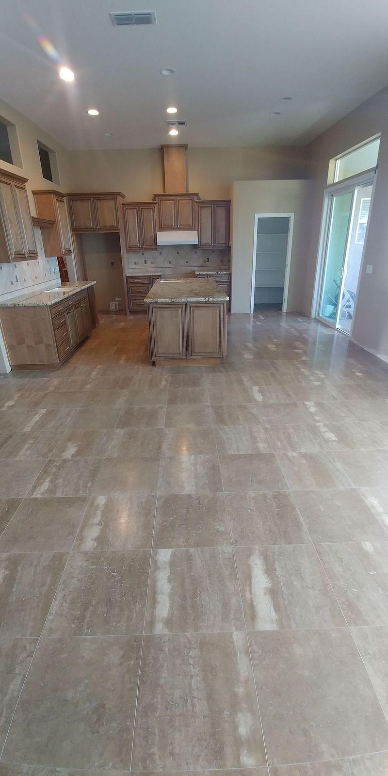 Open kitchen with island, wooden cabinets, and a polished stone floor. Sunlight streams in from a window.