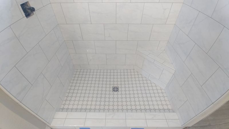 Tiled shower stall in progress with a center drain, white tiles, and grey grout.