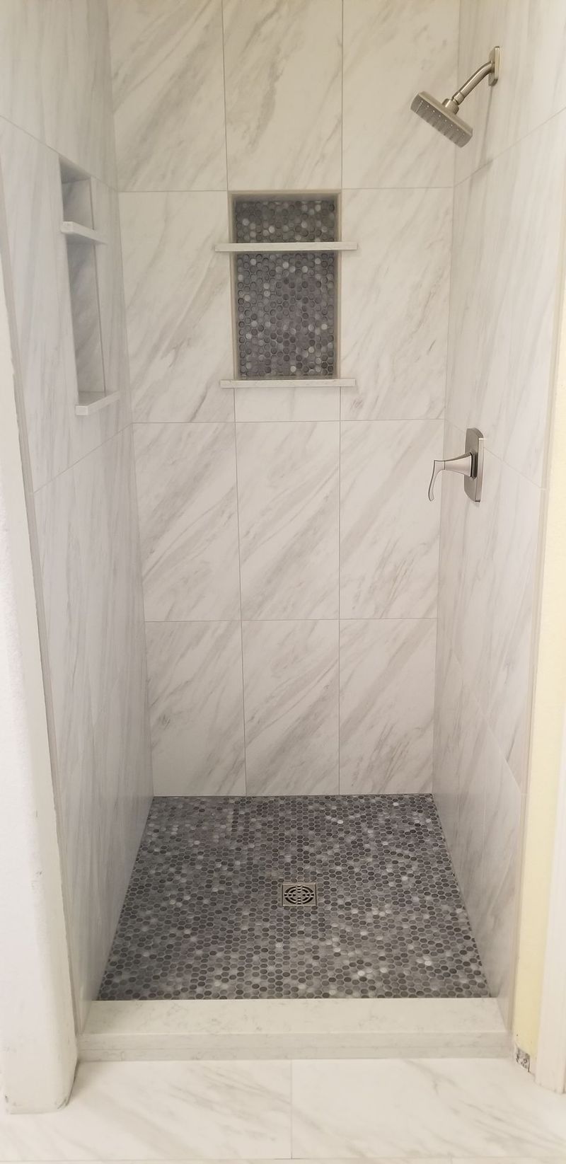 Shower with gray stone floor, white marble-look tile walls, built-in shelving, and silver fixtures.