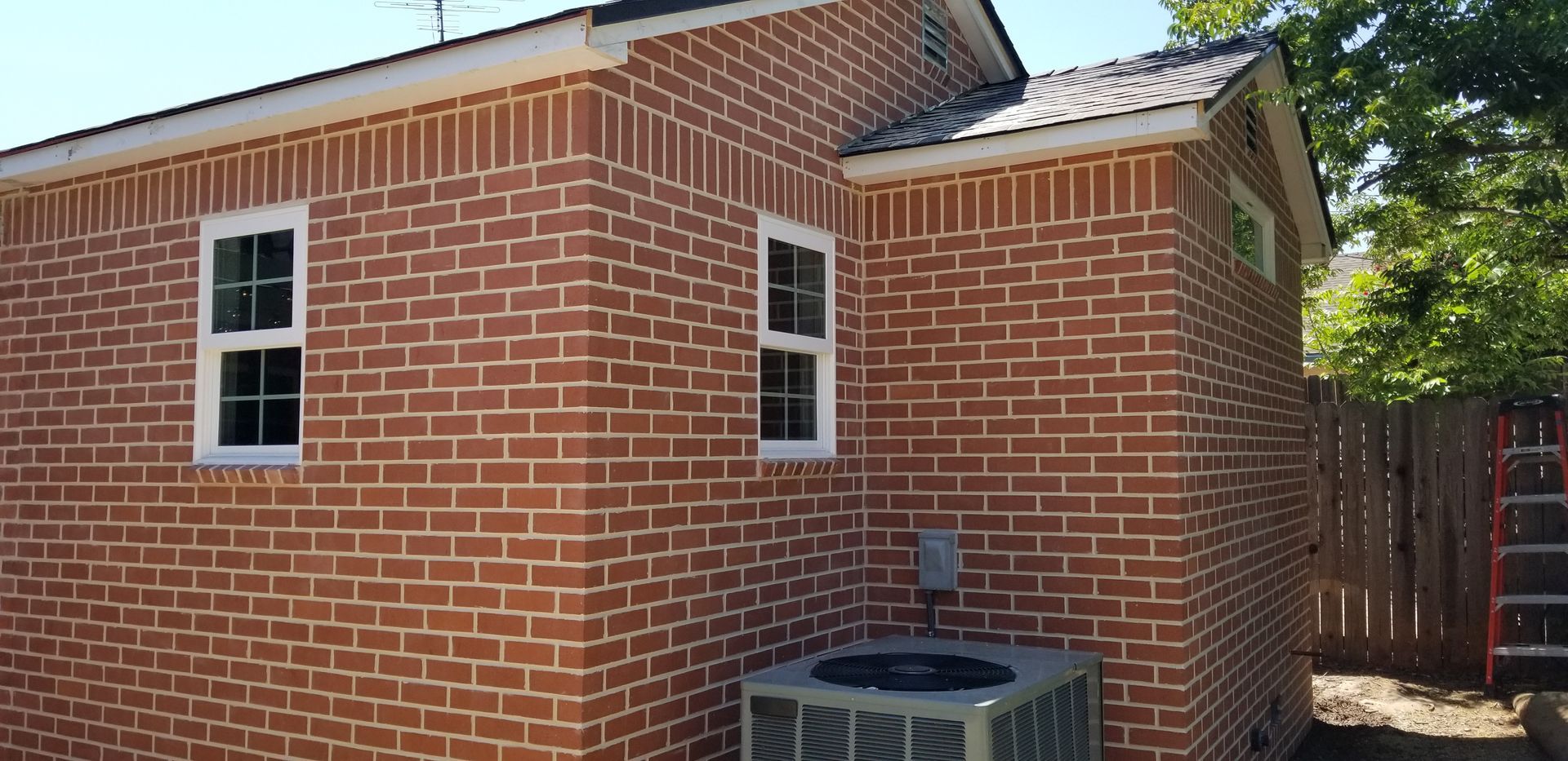 Brick house exterior with two white-framed windows, an air conditioning unit, and a fence.