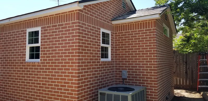Brick house exterior with two white-framed windows, an air conditioning unit, and a fence.