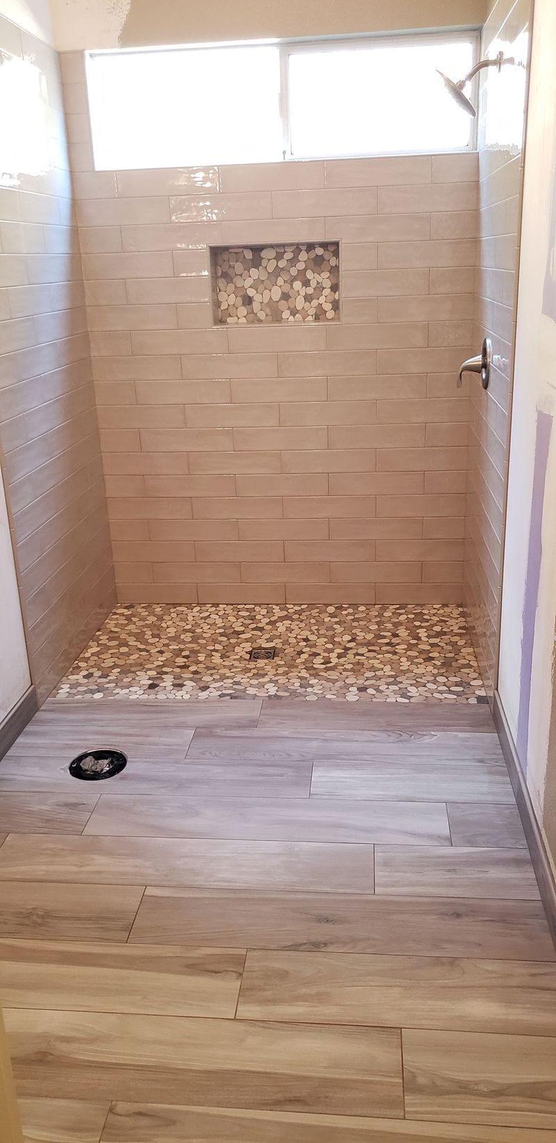 Shower stall with pebble floor and stone tile walls. Wood-look tile floor outside shower. Window above.