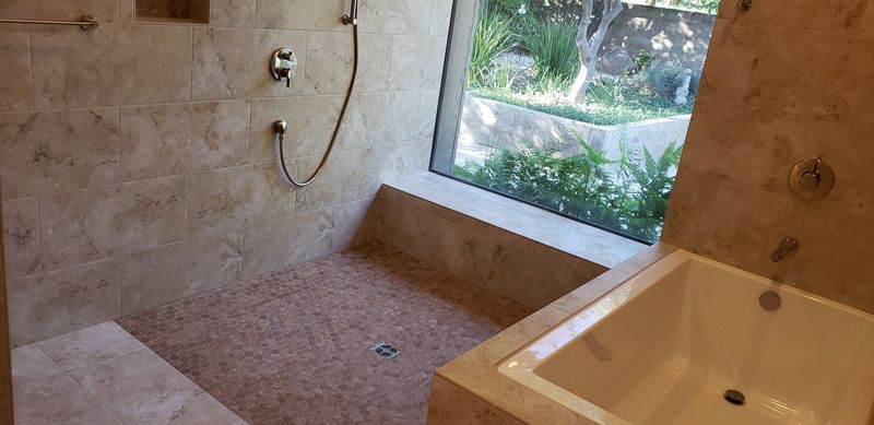 Bathroom with a shower and bathtub. Light brown stone tiles cover the walls and floor. A large window offers a view.