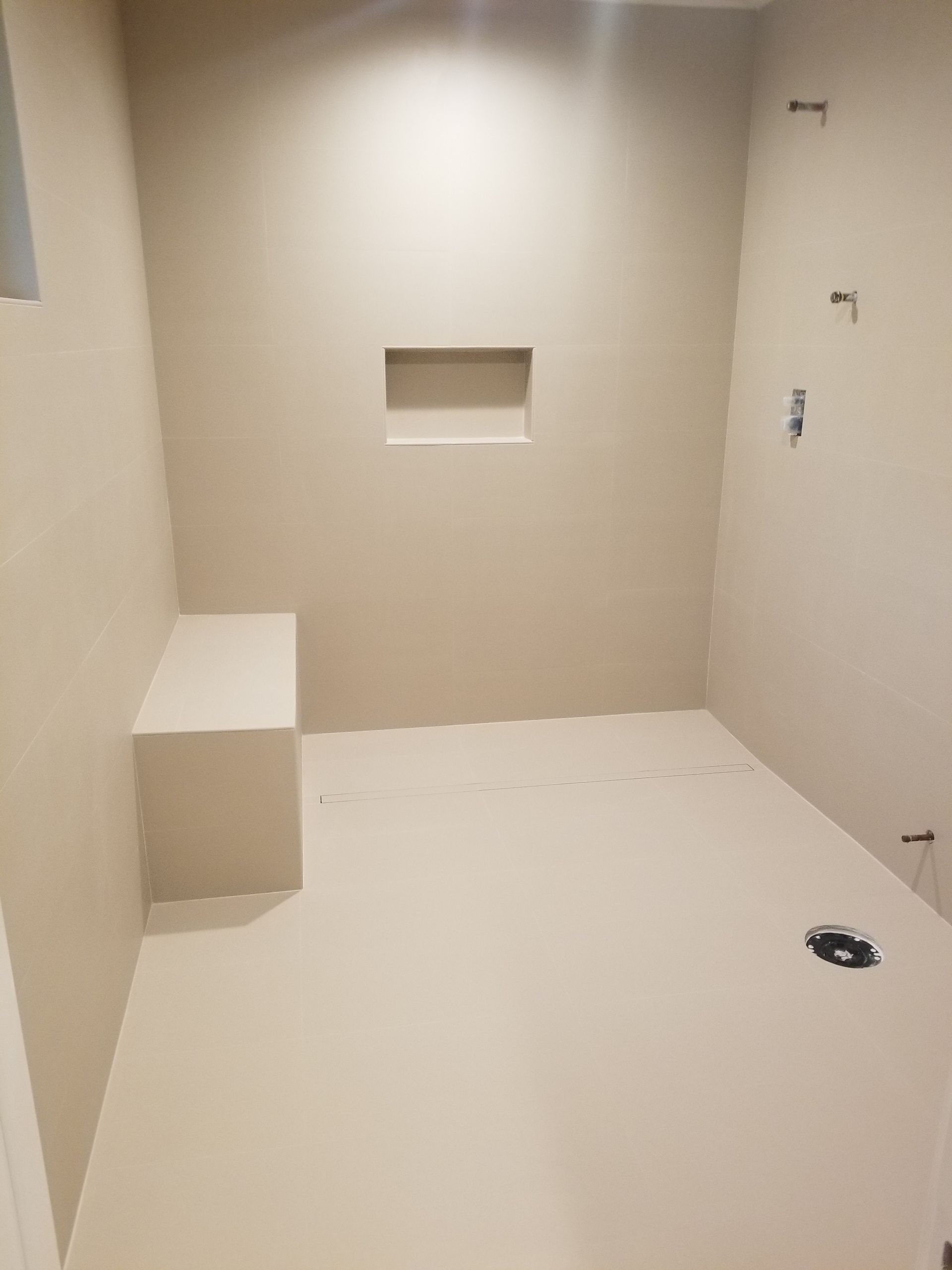 Empty shower with built-in bench and recessed shelf, light beige walls and floor, fixtures installed.