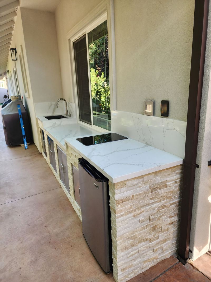 Outdoor kitchen with a white countertop, sink, cooktop, and small refrigerator. Stone-faced base.
