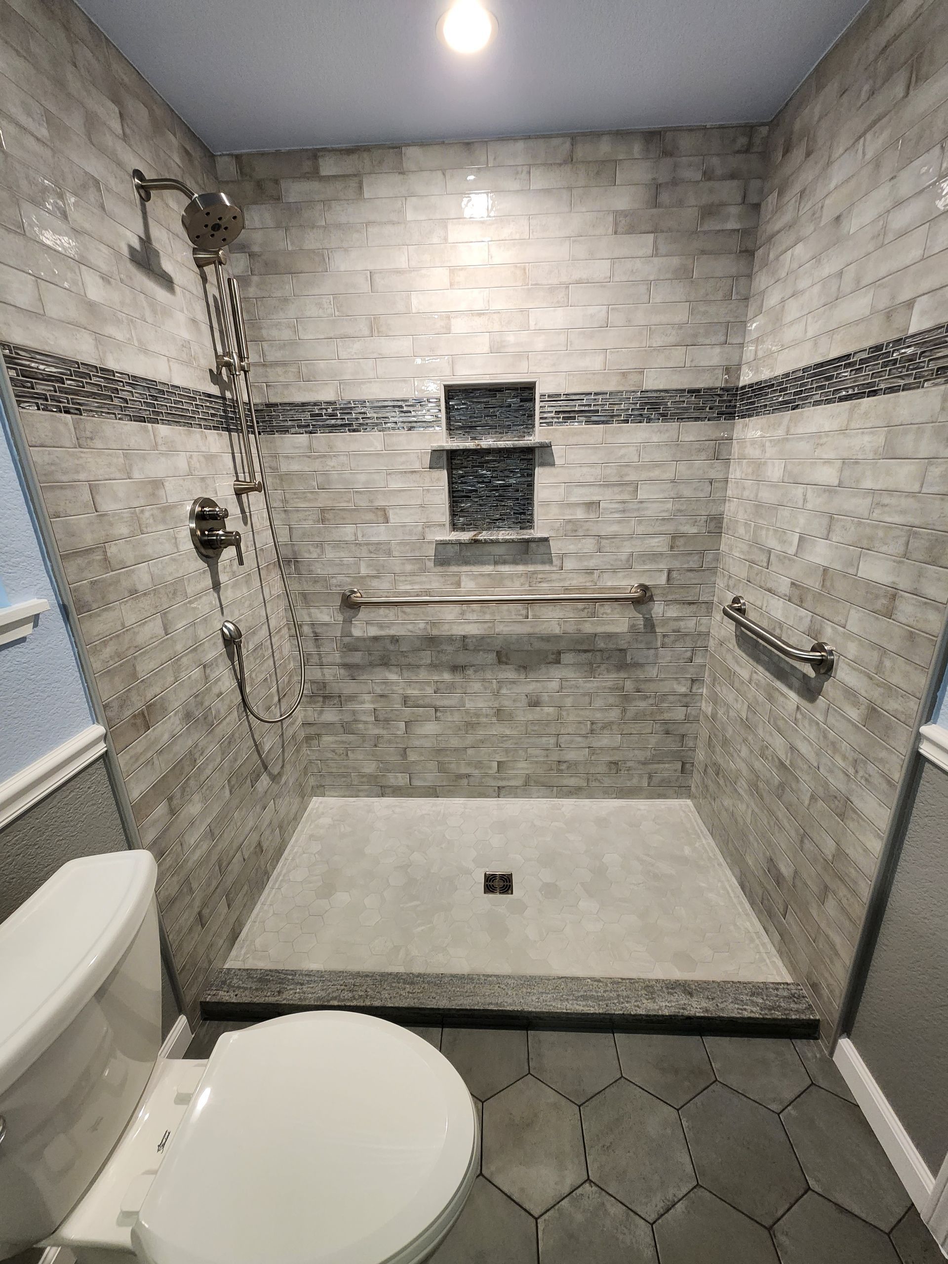 Bathroom with stone tile shower and built-in shelves. Gray tiles, silver fixtures. Toilet in foreground.
