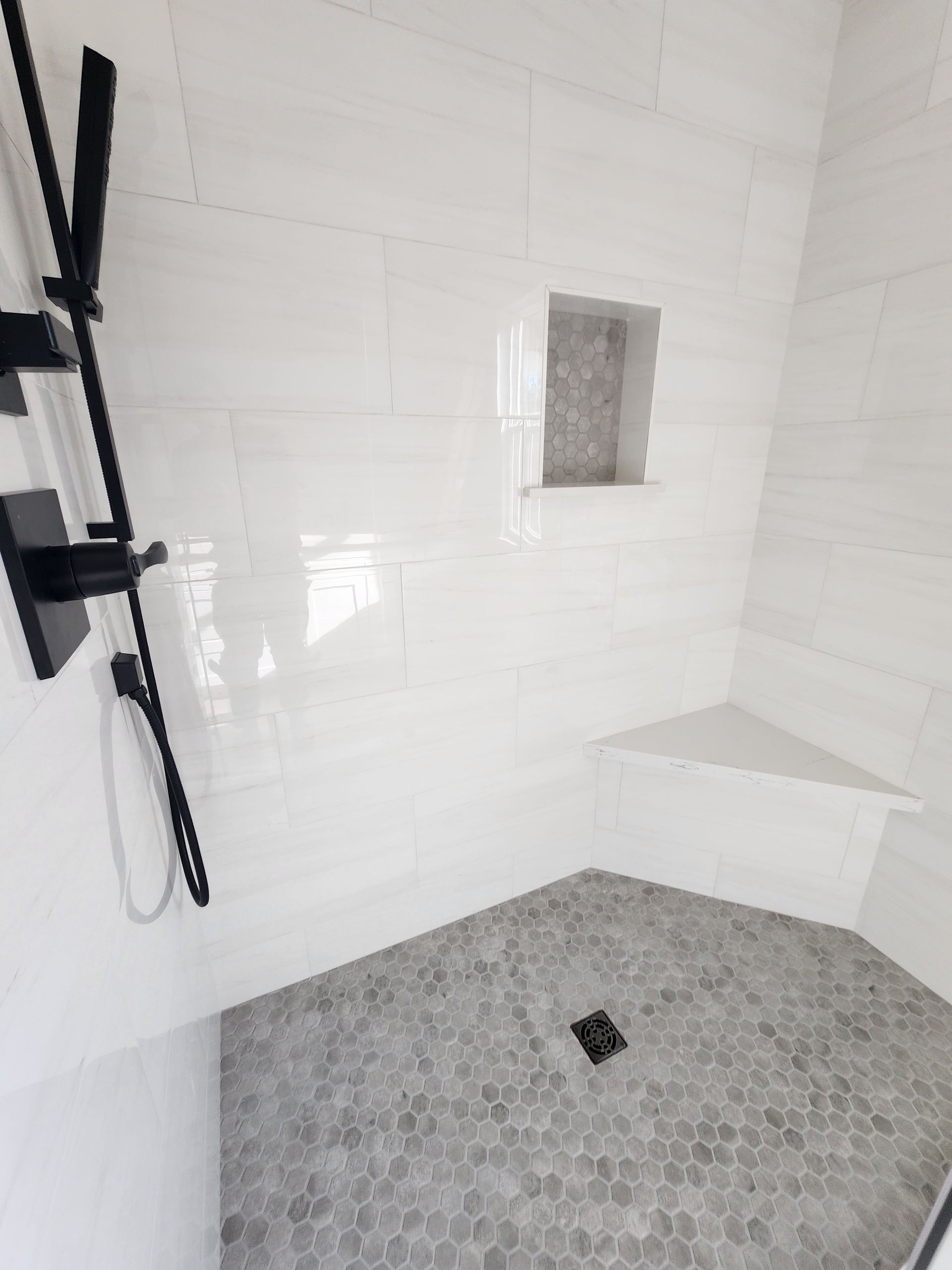 White tiled shower with a gray honeycomb tile floor, a black shower fixture and a built in shelf.