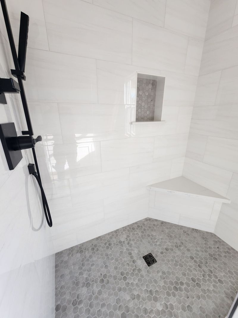 White tiled shower with a gray honeycomb tile floor, a black shower fixture and a built in shelf.
