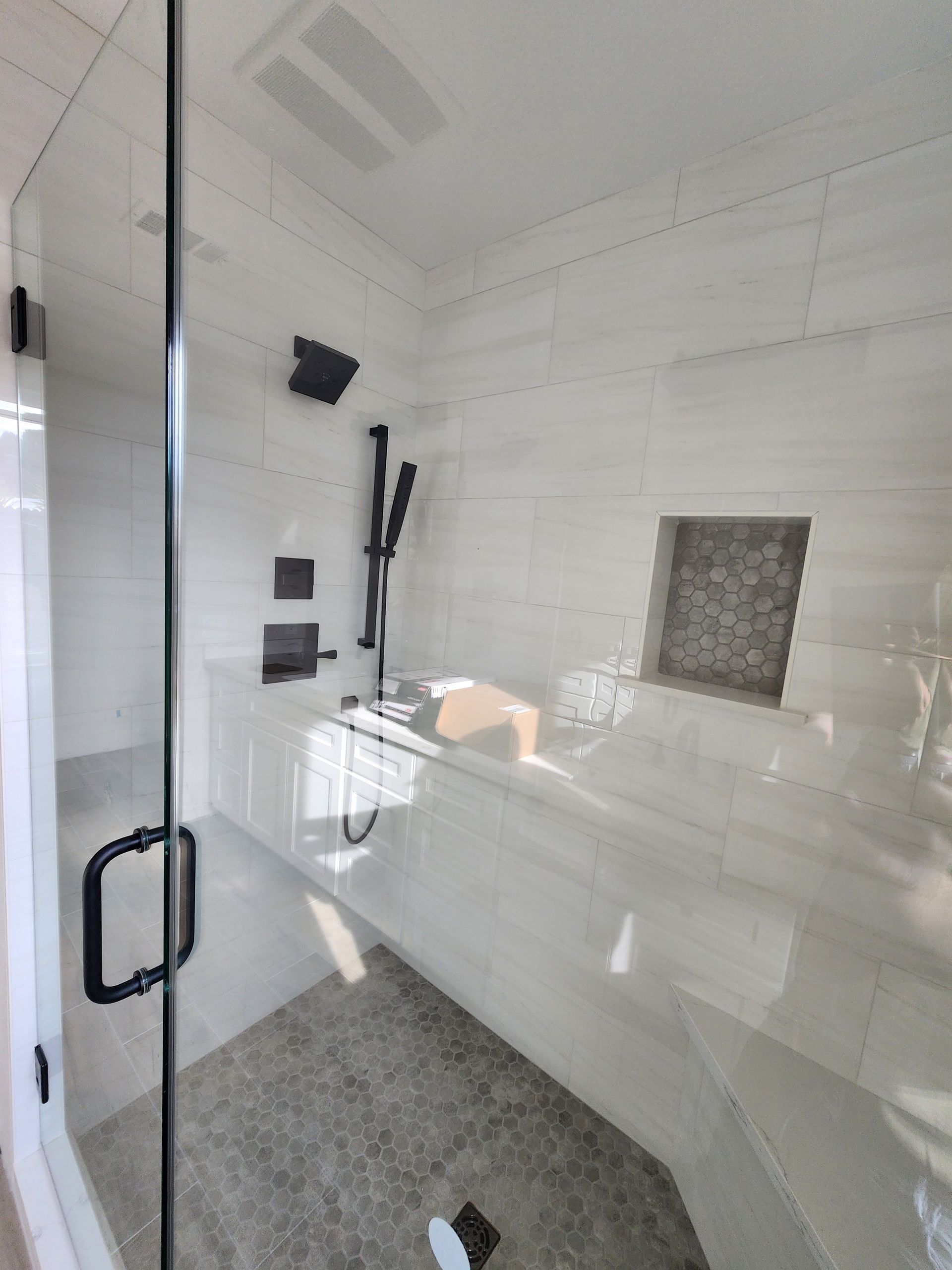 Modern, all-white shower with glass door, black fixtures, built-in bench, and stone tile floor.