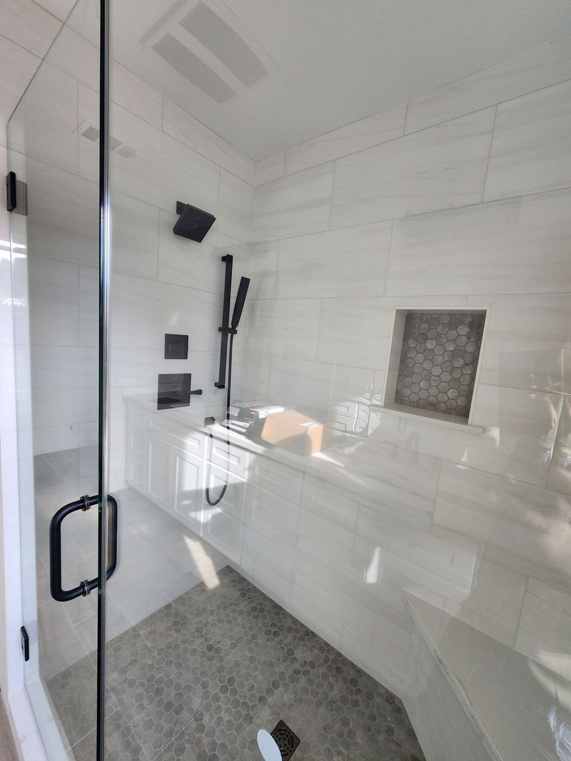 Modern, all-white shower with glass door, black fixtures, built-in bench, and stone tile floor.