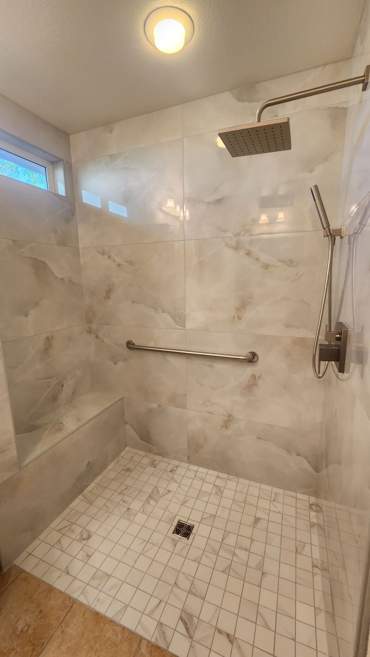Accessible shower with marble-like tile, overhead showerhead, grab bar, and small tiled floor.