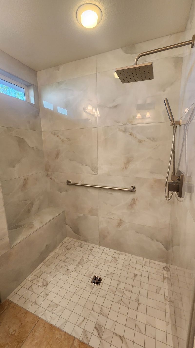 Accessible shower with marble-like tile, overhead showerhead, grab bar, and small tiled floor.