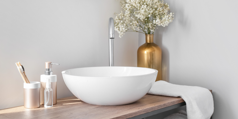 Bathroom sink with white bowl, golden vase with flowers, and toiletries on a wooden shelf.