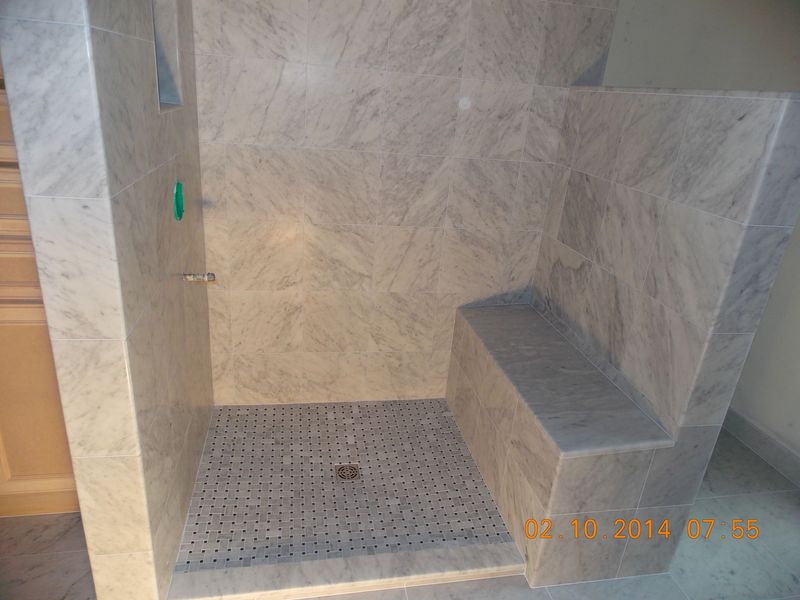 Marble-tiled shower stall with built-in bench. Dark gray tiled floor with central drain. Soap dispenser on wall.