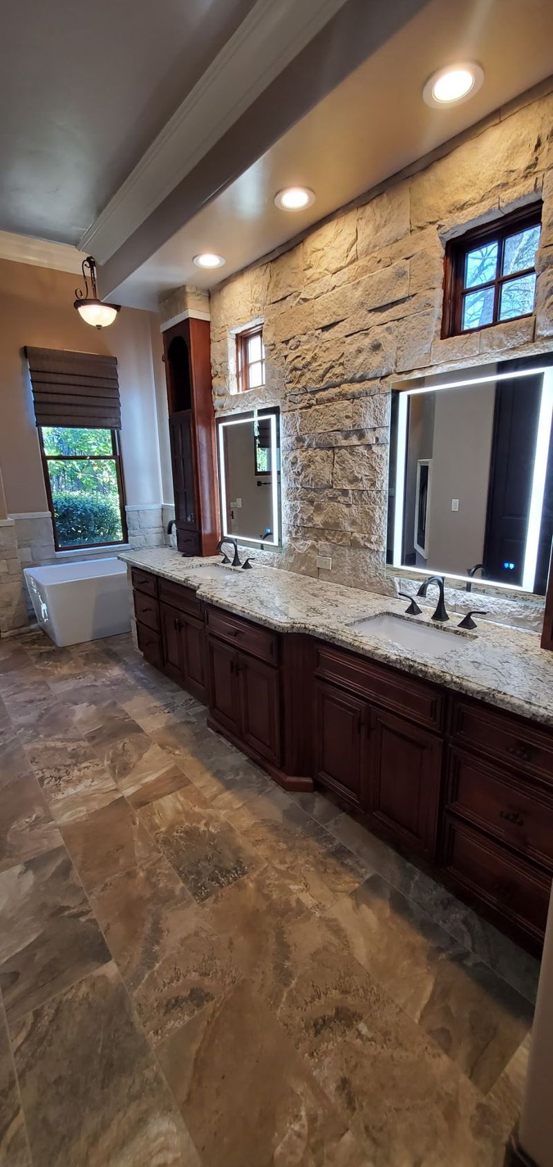Bathroom with stone-textured wall, dark wood vanity, illuminated mirrors, and stone tile floor.