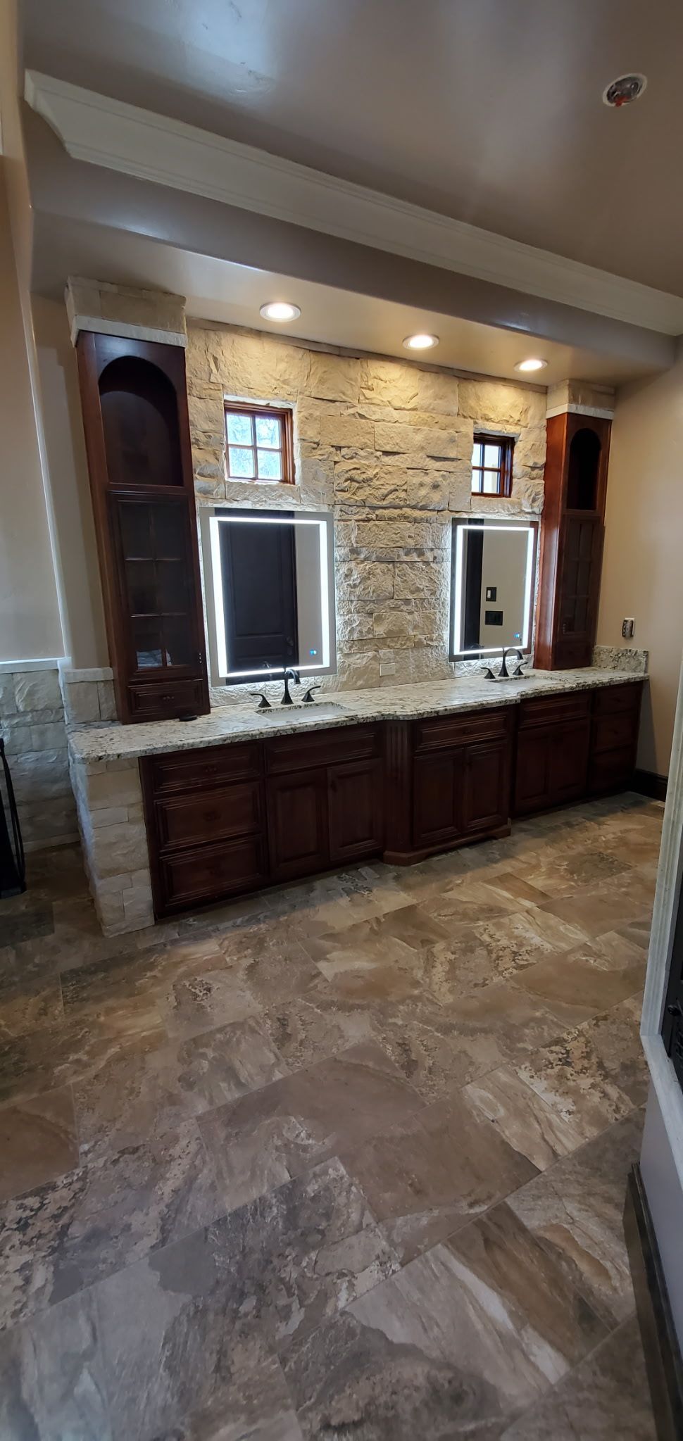 Bathroom with a double vanity. Dark wood cabinets and stone tile. Lighted mirrors and stone backsplash.