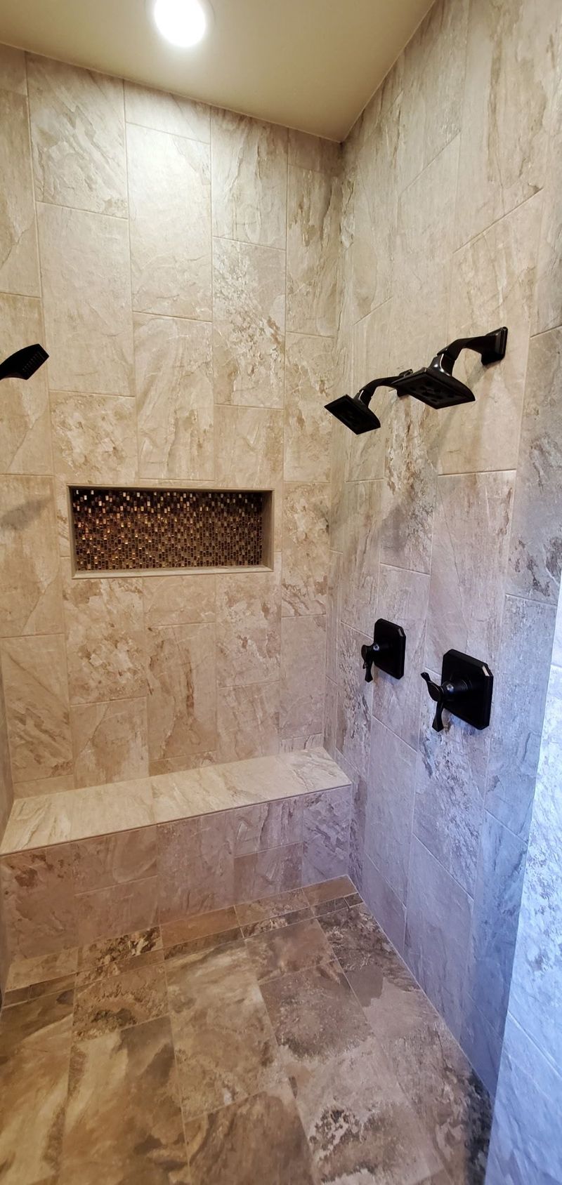 Shower with stone-like tile walls and bench, featuring black showerheads and controls, and a recessed storage niche.