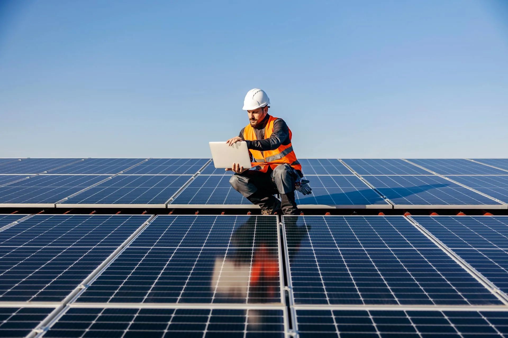 Worker in hard hat inspecting solar panels on a rooftop under a clear blue sky