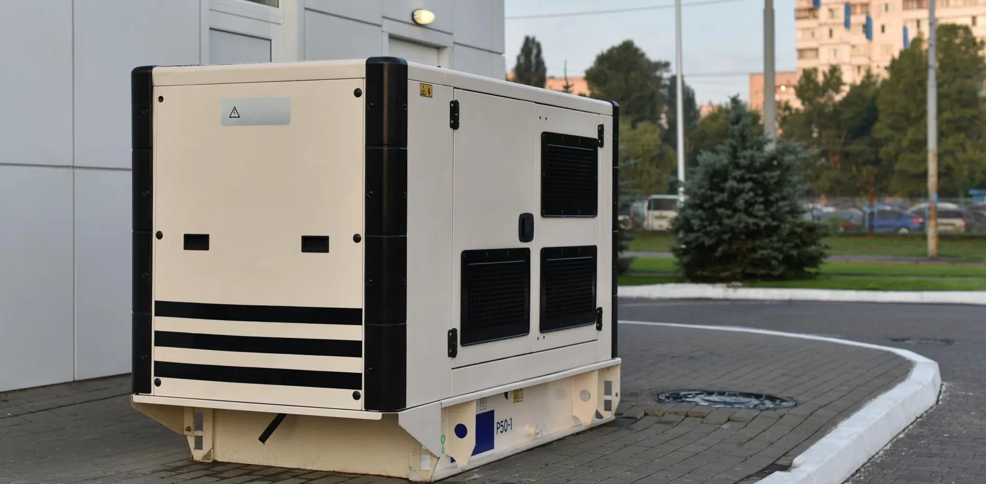 Large beige utility generator on a sidewalk near grass and apartment buildings at dusk