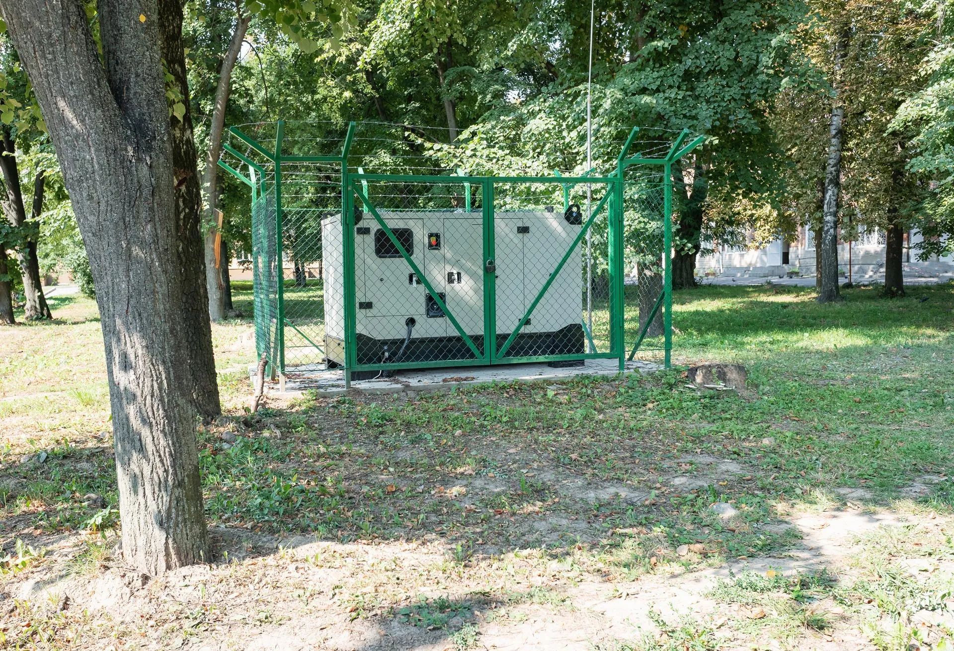 Green fenced utility cabinet in a wooded park area, with trees and grass around it.