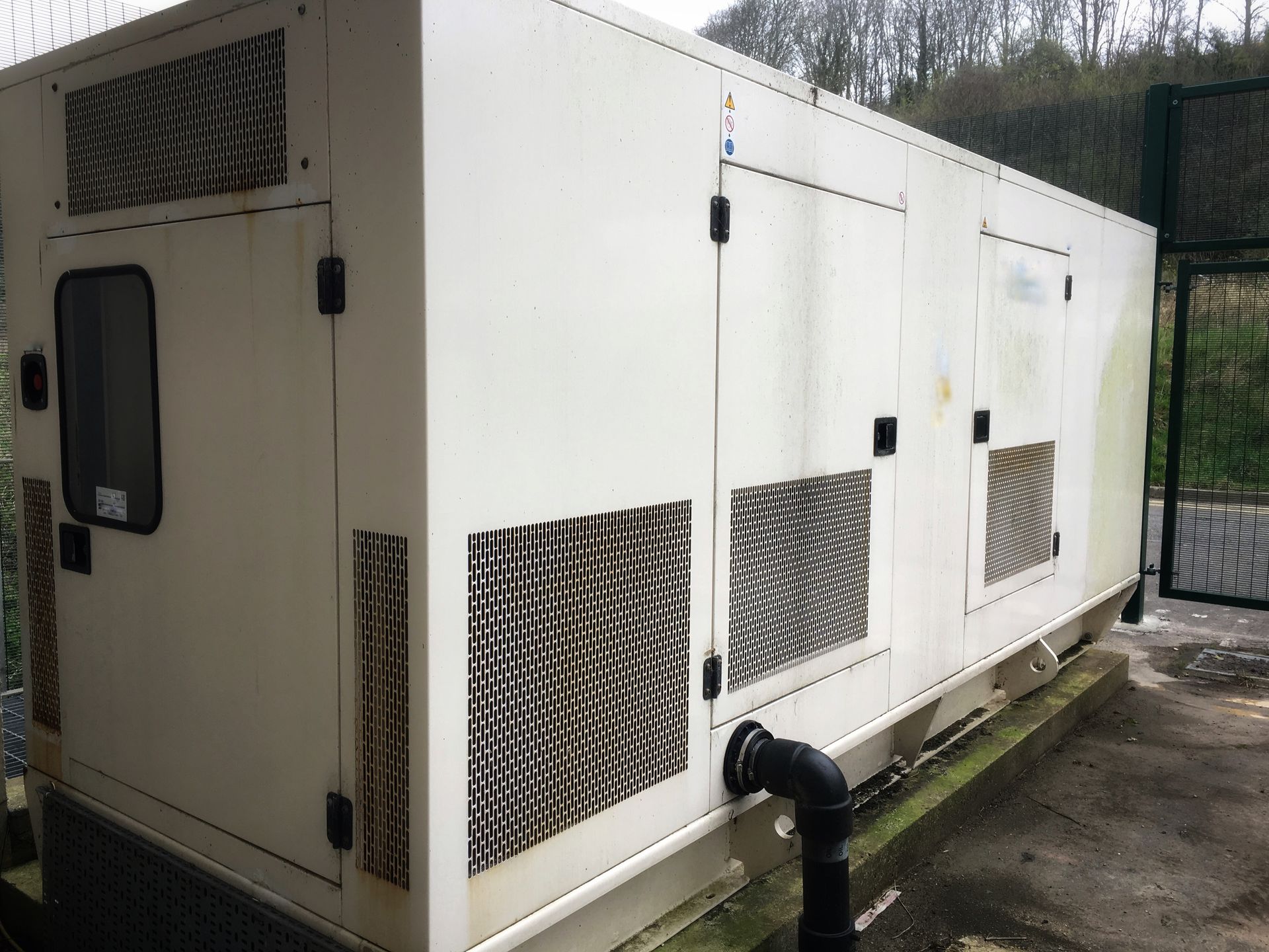 White industrial generator enclosure beside a fence, with vents and a black pipe on a concrete pad