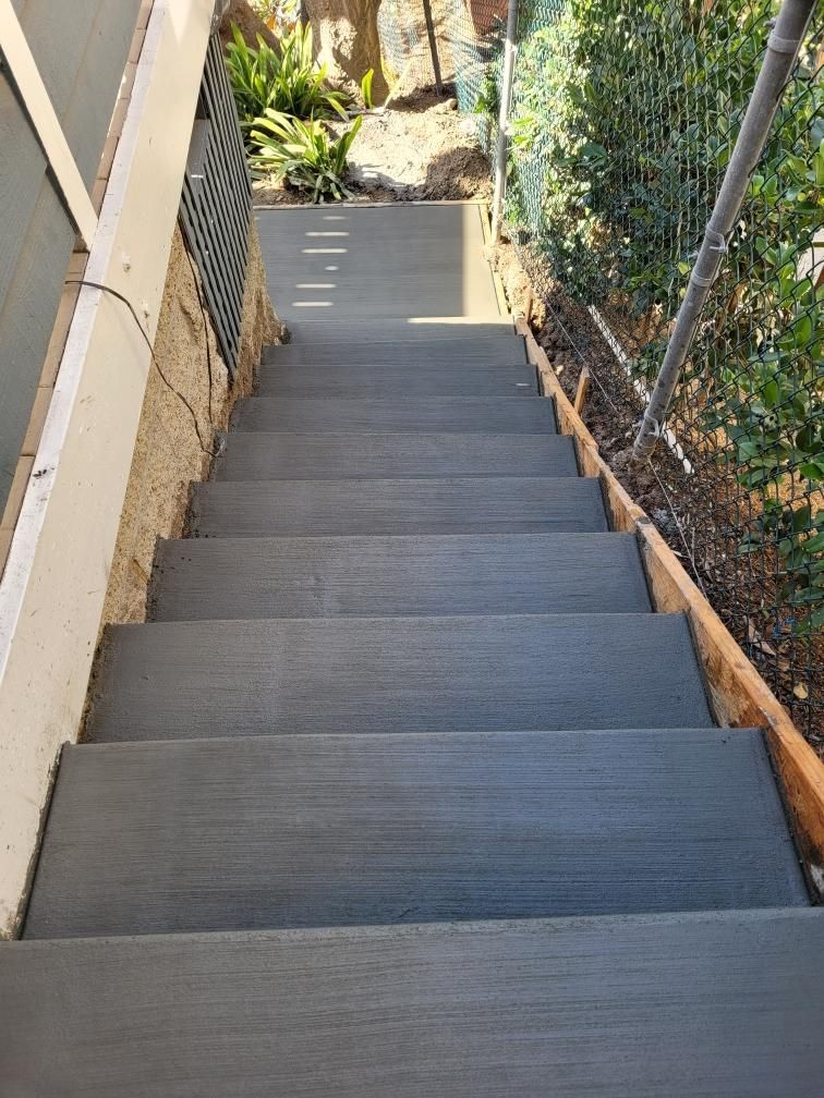 Steep outdoor concrete stairs beside a tan wall and chain-link fence, leading down to a garden path