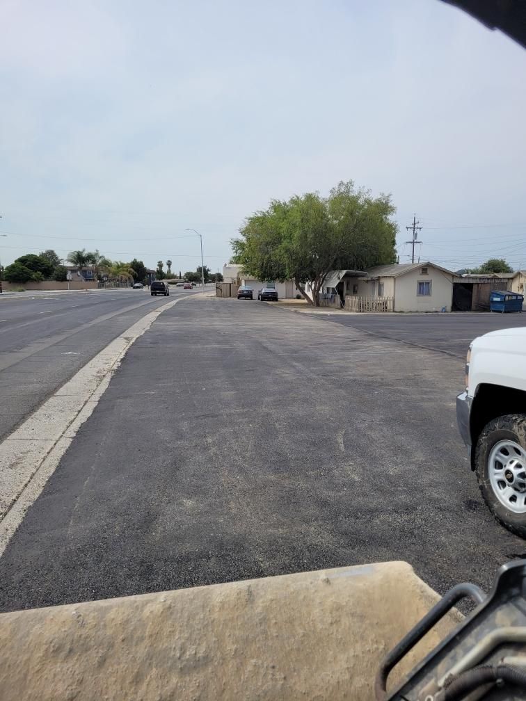 Empty asphalt lot beside a road with houses, trees, and a white truck on the right