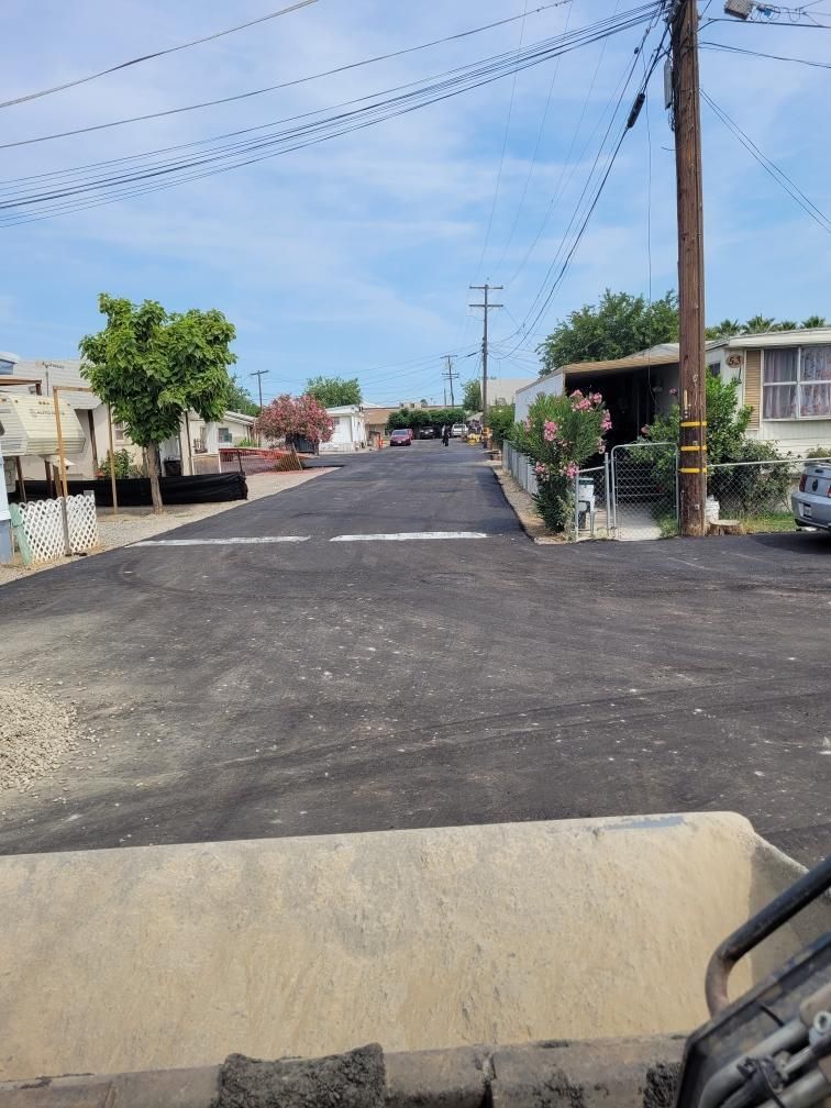 Residential street with fresh black asphalt, beige curbs, houses, trees, and utility poles under a clear blue sky