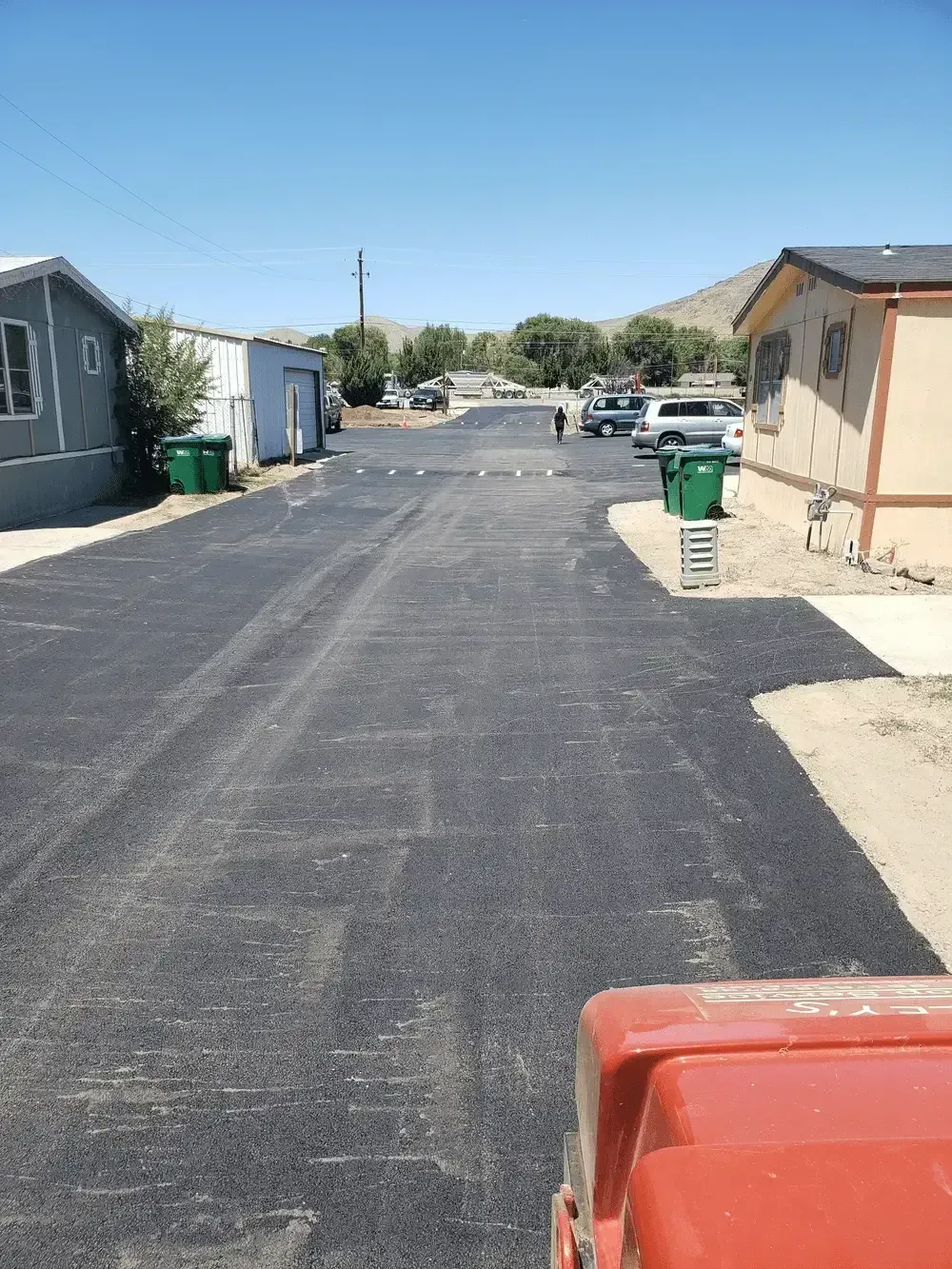 Residential alley with new black pavement, green trash bins, and houses on both sides under a clear sky
