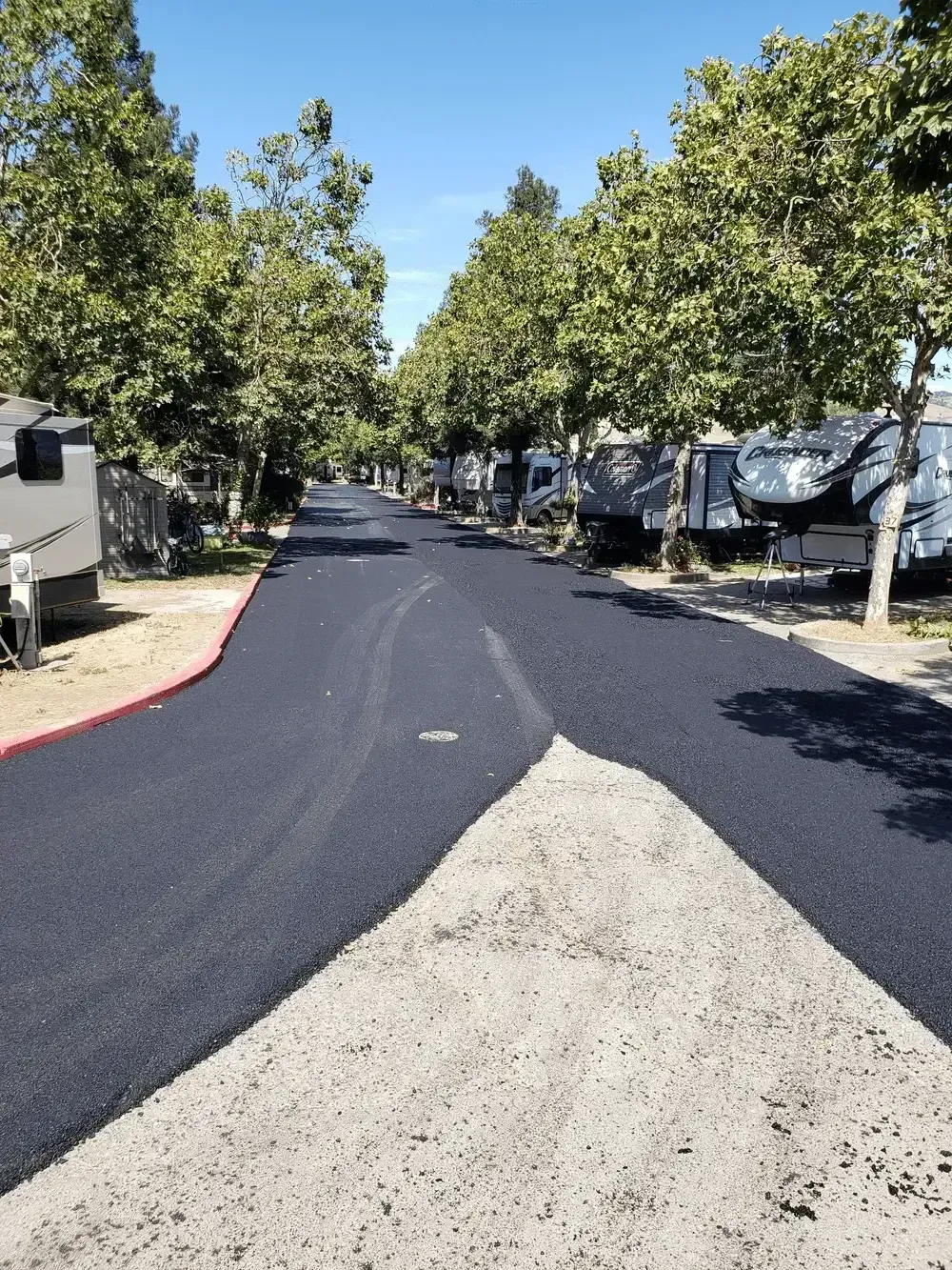 Tree-lined paved road in an RV park, with dark asphalt under repair and trailers on both sides.