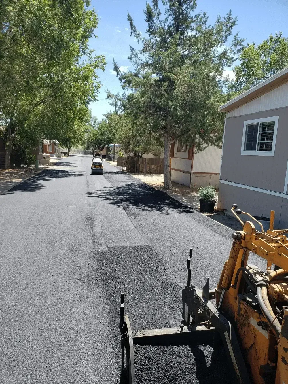 Freshly paved residential street with trees, houses, and roadwork equipment in the foreground