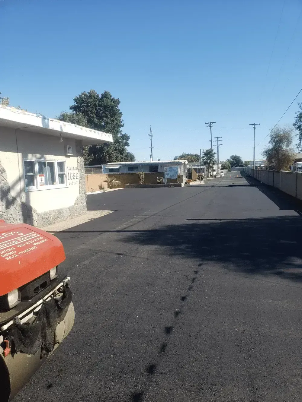 Empty asphalt road beside a white building, with a red vehicle at left and trees under a clear blue sky.