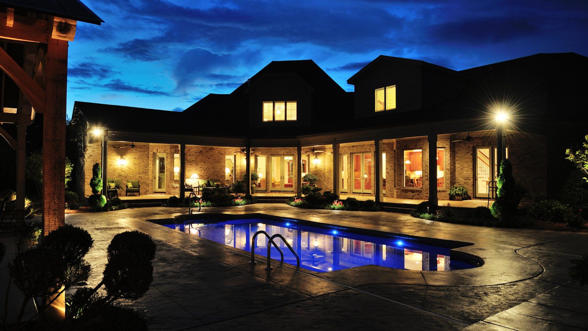 A pool in the backyard of a house.  The picture is taken at night. The house and pool all have outdoor lighting.  The deck around the pool is concrete stamps.