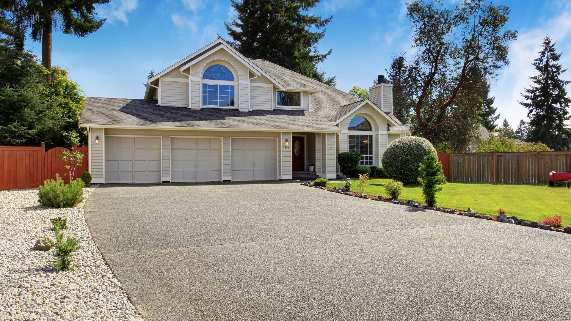 Large concrete driveway leading up to a house with a triple car garage.  The property has nice landscaping with gardens, pebbles and bushes.